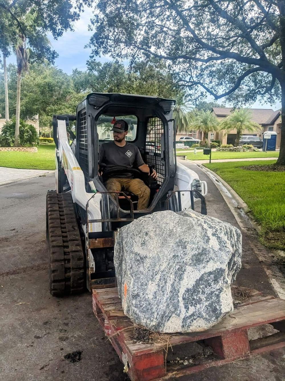 Man operating a skid steer loader with a large rock on a wooden pallet.
