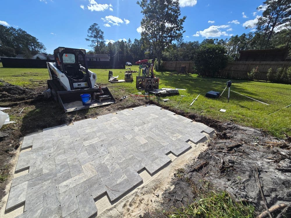 Construction site with a partially laid paver stone patio and a bobcat skid steer nearby.