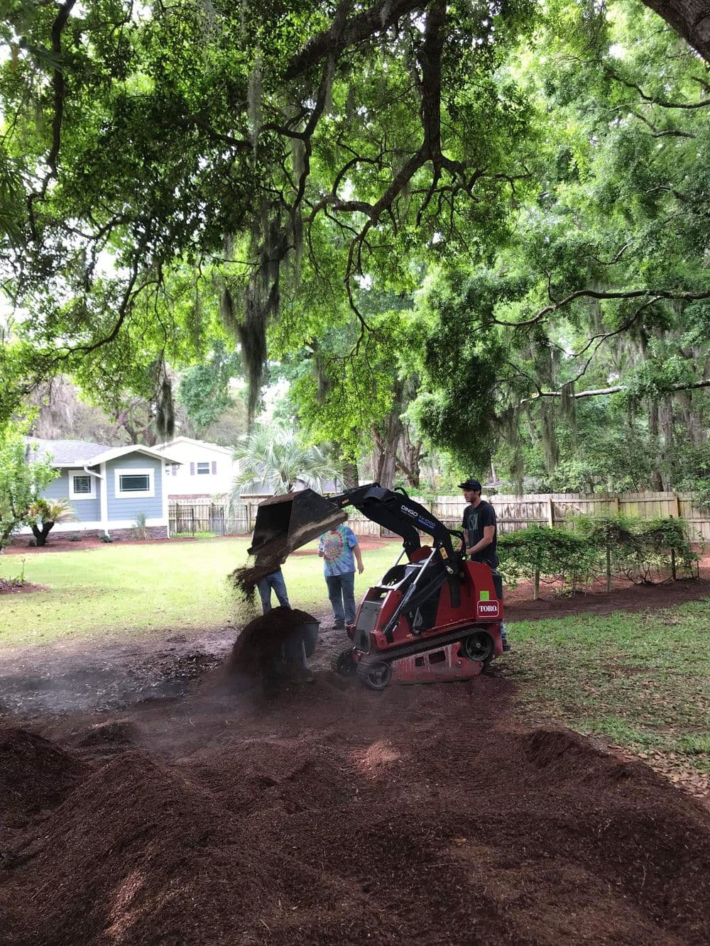 Person using a mini skid steer to move mulch in a residential backyard surrounded by trees.