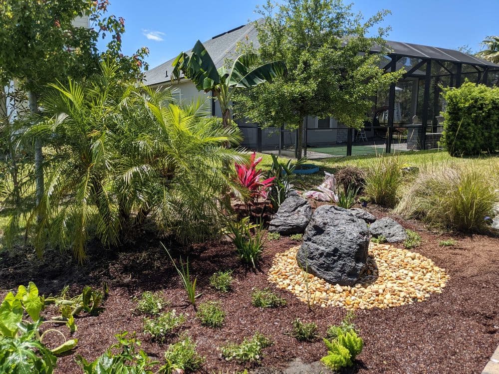 Lush garden landscape with tropical plants, decorative rocks, and pebbled area.