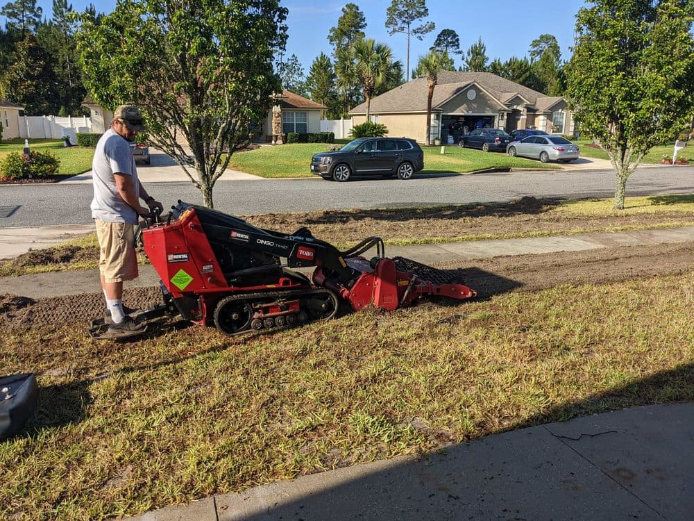 Person operating a mini skid steer during landscaping on a suburban street.