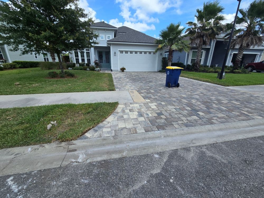 Residential home with paver driveway, blue trash bin, and palm trees under a partly cloudy sky.